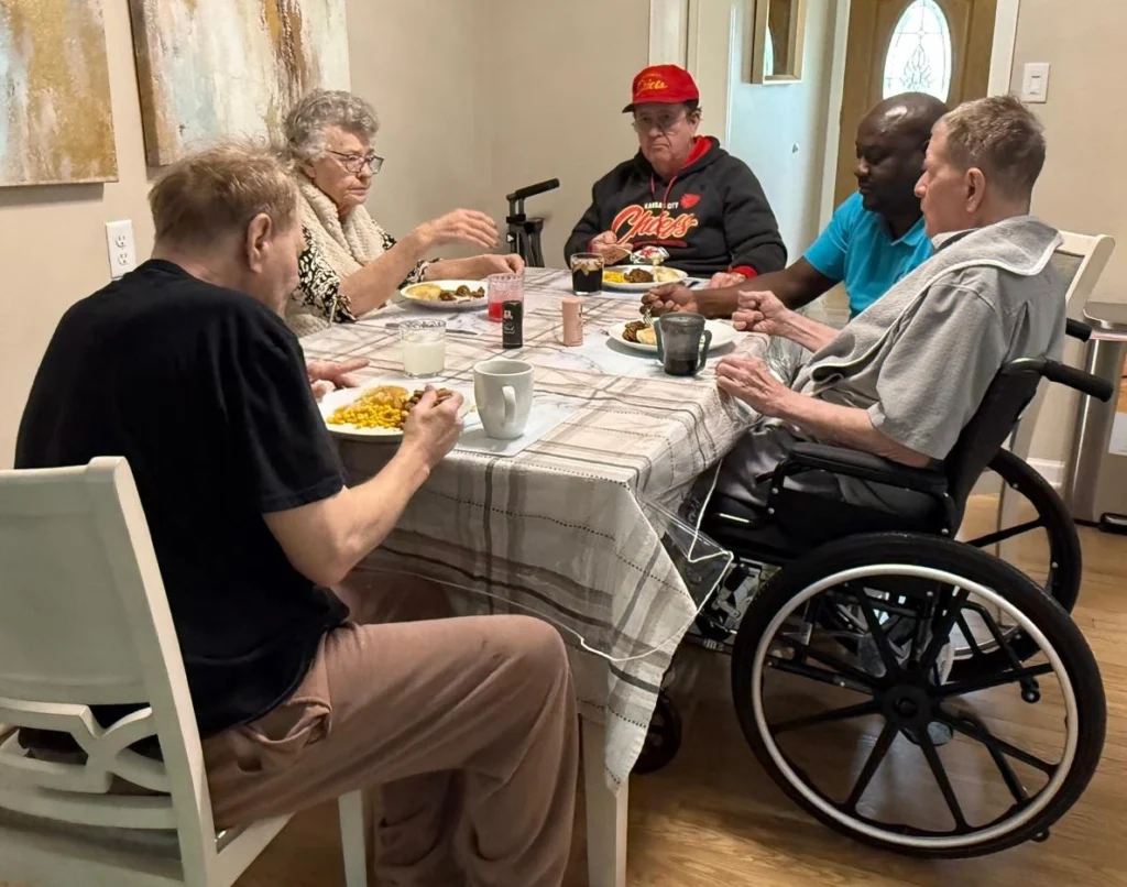 Caregiver Sharing A Meal With Senior Residents At The Dinner Table In A Home Care Setting 1024x806