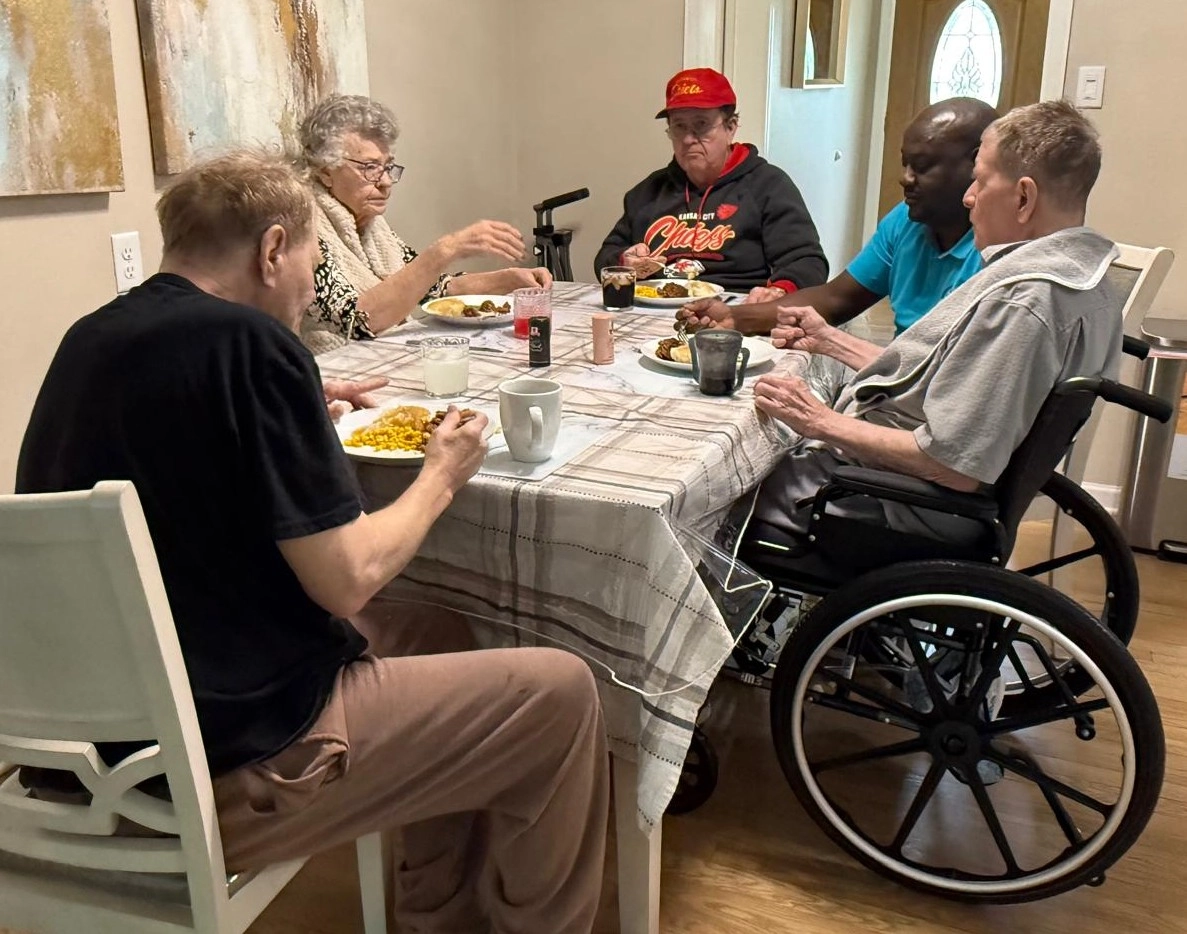 Caregiver Sharing A Meal With Senior Residents At The Dinner Table In A Home Care Setting